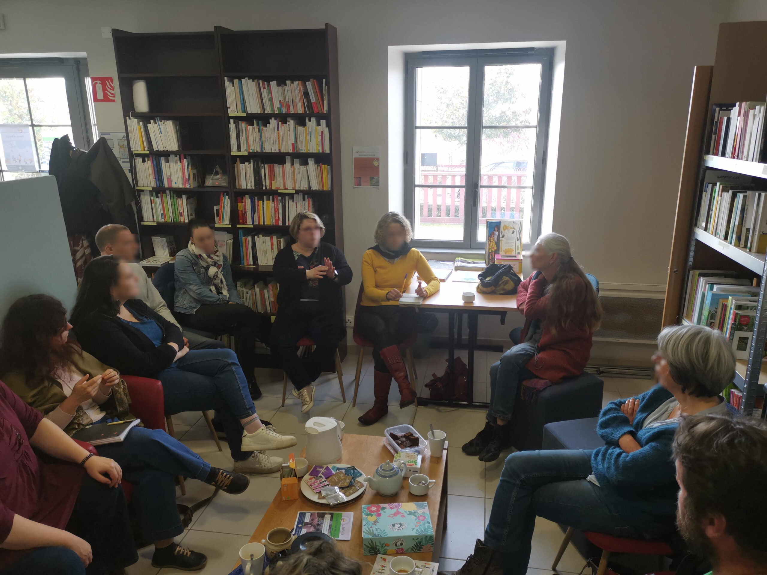 Photo du café des indépendants. 8 personnes autour d'une table avec café et tisanes. En fond des bibliothèques remplies de livres.