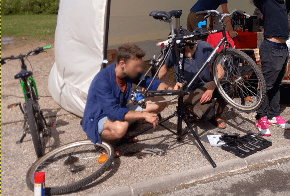 Photo du répare-vélo : on voit une personne en train de démonter un vélo, dont la roue arrière est déjà démontée. Le vélo est porté par un pied d'atelier. E arrière-plan, on voit le barnum et des bénévoles.